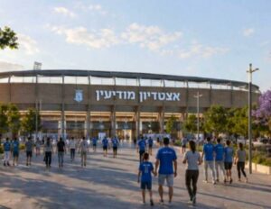 הנחת אבן פינה לקריית הספורט העירונית 28/04/2026 י"א אייר תשפ"ו דף הבית העירייה חדשות העיר הנחת אבן פינה לקריית הספורט העירונית ביום ראשון 26.4, התקיים טקס הנחת אבן פינה לפרויקט הענק של הקמת אצטדיון עירוני מתקדם לכדורגל. הטקס התקיים בהשתתפות ראש העיר חיים ביבס הסגנים, אור גרינפלד, עמיעד טאוב, שלמה פסי ואלעד שמעונוביץ, המשמש גם כמחזיק תיק הספורט, חברי מועצת העיר שמעון גואטה, גלעד ביהרי, איתי בר אלמוג ורן שגב, נציגי הנהלת עירוני מודיעין, מאמן עירוני מודיעין ארז בנודיס ומייסד המועדון, דוד בן שימול. נציגי העירייה והחברה הכלכלית לפיתוח מודיעין. המהלך, בהובלת ראש העיר חיים ביבס, מגיע לאחר עבודת מטה מקצועית, שכללה בדיקות שטח, למידת תהליכים בערים אחרות, וקידום פתרונות תחבורתיים ותכנוניים שישרתו את האצטדיון ואת הציבור הרחב. הפרויקט, בהובלת ראש העיר חיים ביבס יוצא לדרך במרכז עינב, במיקום אסטרטגי המציע נגישות תחבורתית מצוינת בזכות תחנת הרכבת פאתי מודיעין הסמוכה וחניון של כ-750 מקומות. האצטדיון יכיל כ־6,000 מושבים, ויעמוד בכל התקנים והדרישות של ההתאחדות לכדורגל בישראל לקיום משחקים בליגה הלאומית ובהמשך גם בליגת העל. המתקן ישמש גם לאירועי תרבות גדולים. העלות הכוללת של הקמת האצטדיון, כולל עבודות הפיתוח, נאמדת בכ־150 מיליון שקלים. כדי להאיץ את התהליך, תבוצע הבנייה בשיטת "תכנון וביצוע", שתאפשר עמידה בלוחות זמנים צפופים תוך שמירה על סטנדרטים גבוהים. לצד האצטדיון, יוקם גם היכל ספורט רב תכליתי חדש, ובו כ־2,600 מקומות ישיבה, אשר יוכל לארח משחקי כדורסל, כדוריד ואירועי תרבות נוספים, מהלך שיחזק את מעמדה של מודיעין מכבים רעות כמרכז ספורט אזורי משמעותי. כך ייראה האצטדיון החדש (הדמייה באמצעות AI) באדיבות דוברות העיריי